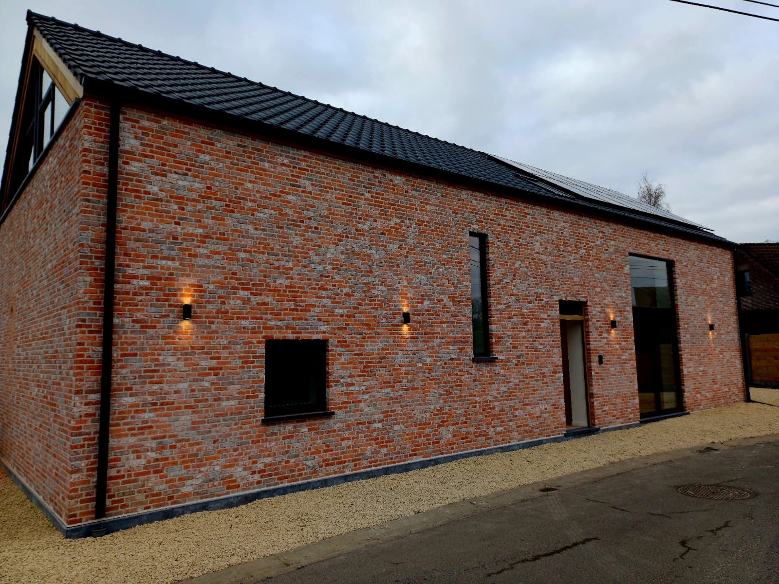 Elewacja budynku z czerwonej cegły z czarnym dachem i oknami, oświetlona zewnętrznymi lampami ściennymi, z panelami słonecznymi na dachu. Widok z poziomu ulicy.