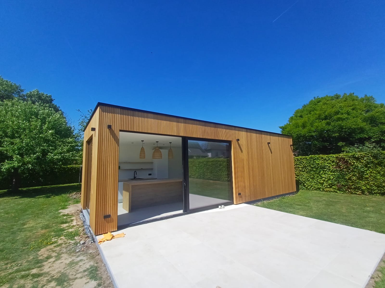 Nowoczesny, drewniany pawilon ogrodowy z przesuwnymi szklanymi drzwiami i zewnętrznym tarasem z jasnych płyt. Widok na minimalistyczne wnętrze z wyspą kuchenną.