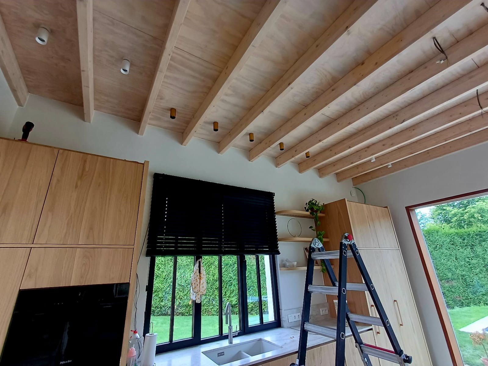 Nowoczesna kuchnia z drewnianym sufitem rastrowym i punktowym oświetleniem. Widoczna drabina, okno z czarnymi żaluzjami i fragment zabudowy kuchennej.