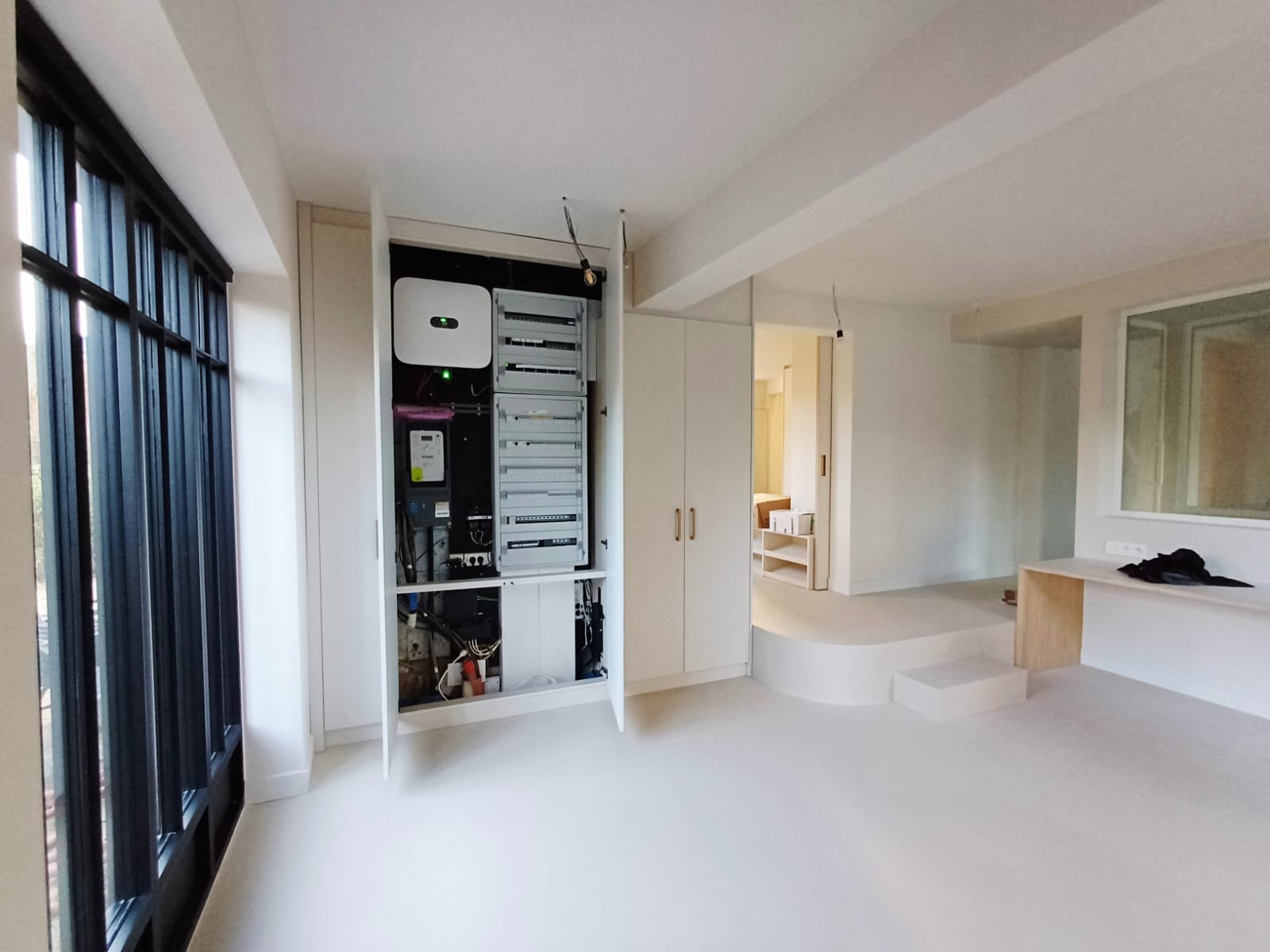 Nowoczesna instalacja elektryczna w szafie wnękowej, widoczny inwerter i rozdzielnica w minimalistycznym wnętrzu z dużym oknem i podwyższoną podłogą.