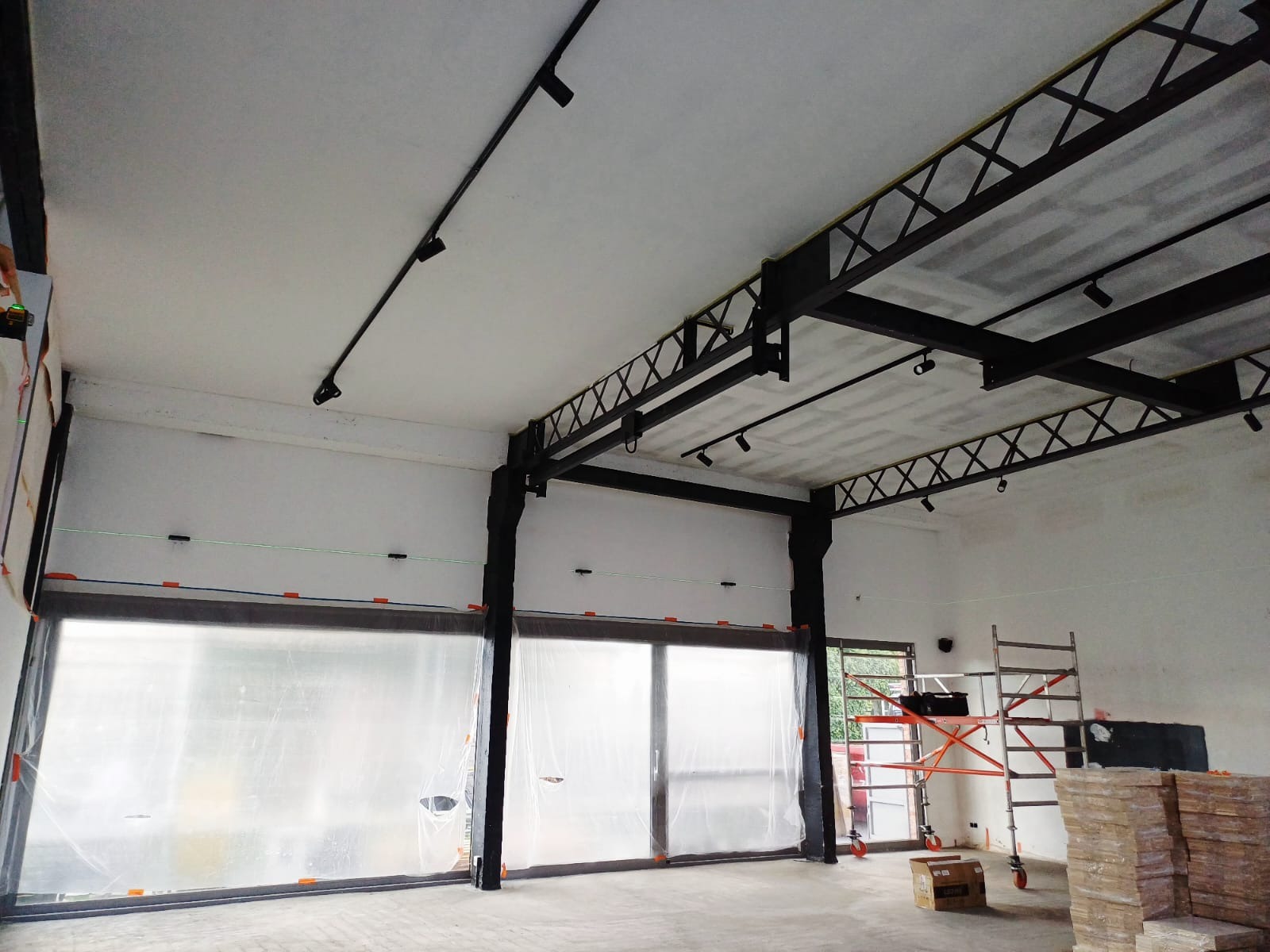 Wnętrze w trakcie montażu oświetlenia szynowego, widoczne czarne szyny i reflektory na suficie, okna zabezpieczone folią, rusztowanie i materiały budowlane. Laser krzyżowy w użyciu.