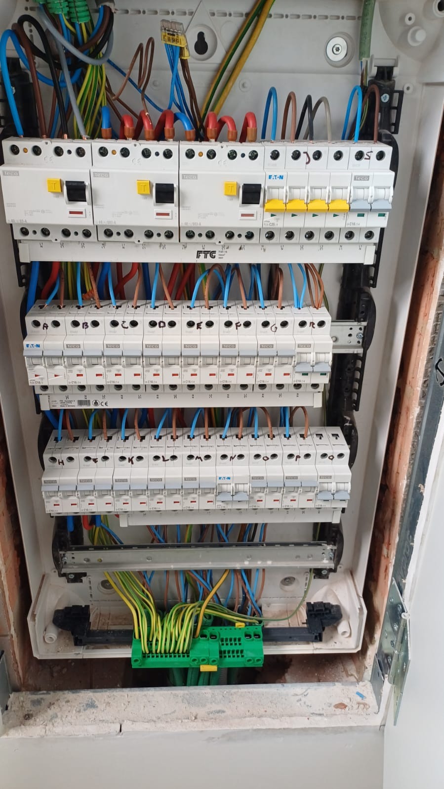 Rozdzielnia elektryczna w trakcie montażu. Widoczne bezpieczniki, przewody w różnych kolorach i złącza. Biała obudowa, surowe wykończenie ściany.