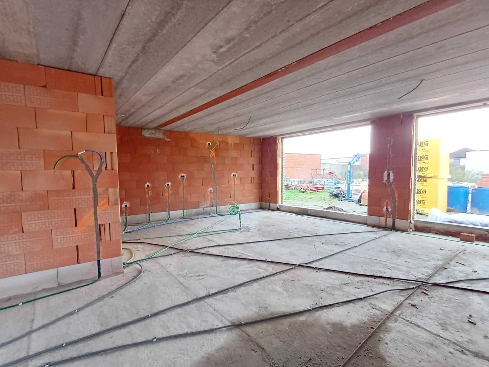 Surowy stan wnętrza budynku w trakcie budowy: widoczne instalacje elektryczne i wodne w ścianach z cegły, ułożone przewody w posadzce, betonowy strop.