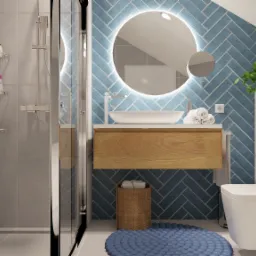 Nowoczesna łazienka z niebieskimi płytkami ułożonymi w jodełkę, okrągłym lustrem z podświetleniem LED, drewnianą szafką pod umywalką i toaletą podwieszaną.