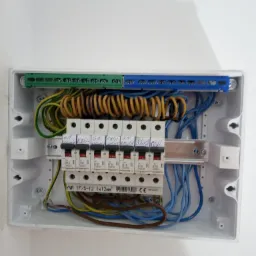 Otwarta skrzynka rozdzielcza z bezpiecznikami i przewodami w różnych kolorach, zamontowana na białej ścianie, widoczne opisy bezpieczników pisane odręcznie.