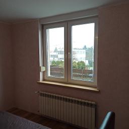 Zaklad ogólnobudowlany Gniewko Surdyk - Pokój po odświeżeniu z widokiem na ogród przez okno, ściany pokryte jasnoróżową tapetą, pod oknem grzejnik, w rogu widoczne rolki tapet.