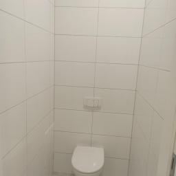 Zaklad ogólnobudowlany Gniewko Surdyk - Wykończona toaleta z białymi, kwadratowymi płytkami na ścianach, podwieszana miska WC i szarymi fugami, widoczny fragment drzwi po prawej stronie.
