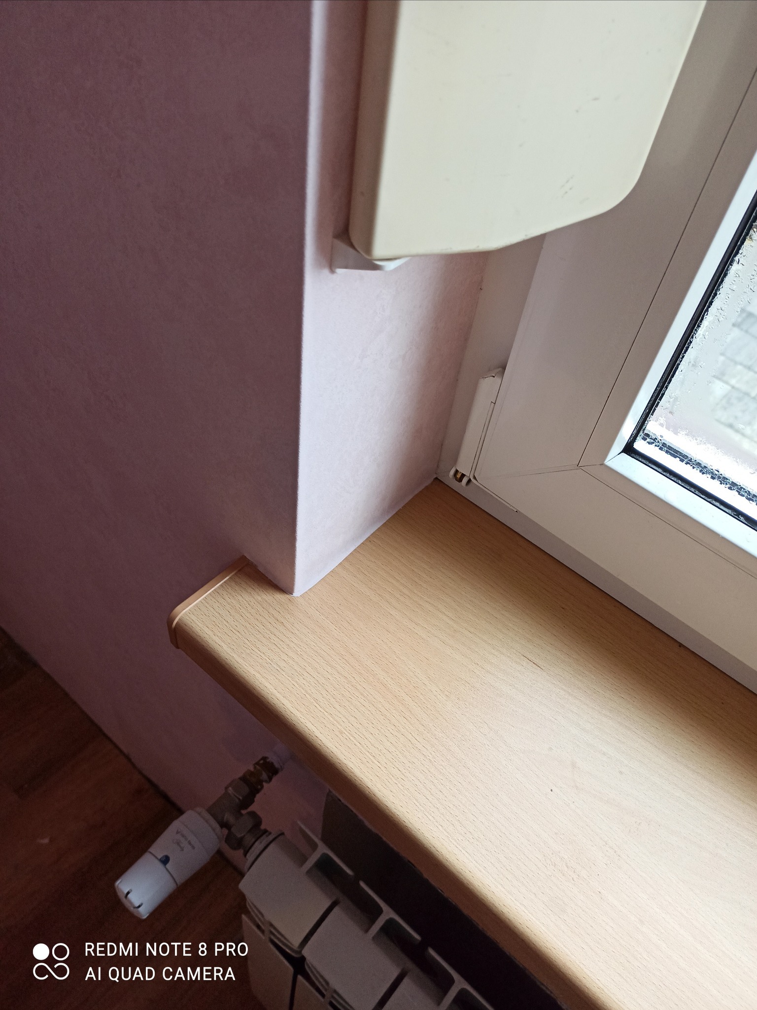 Fragment pokoju z różową ścianą, jasnym parapetem i grzejnikiem z termostatem, widoczne krople wody na szybie okna.