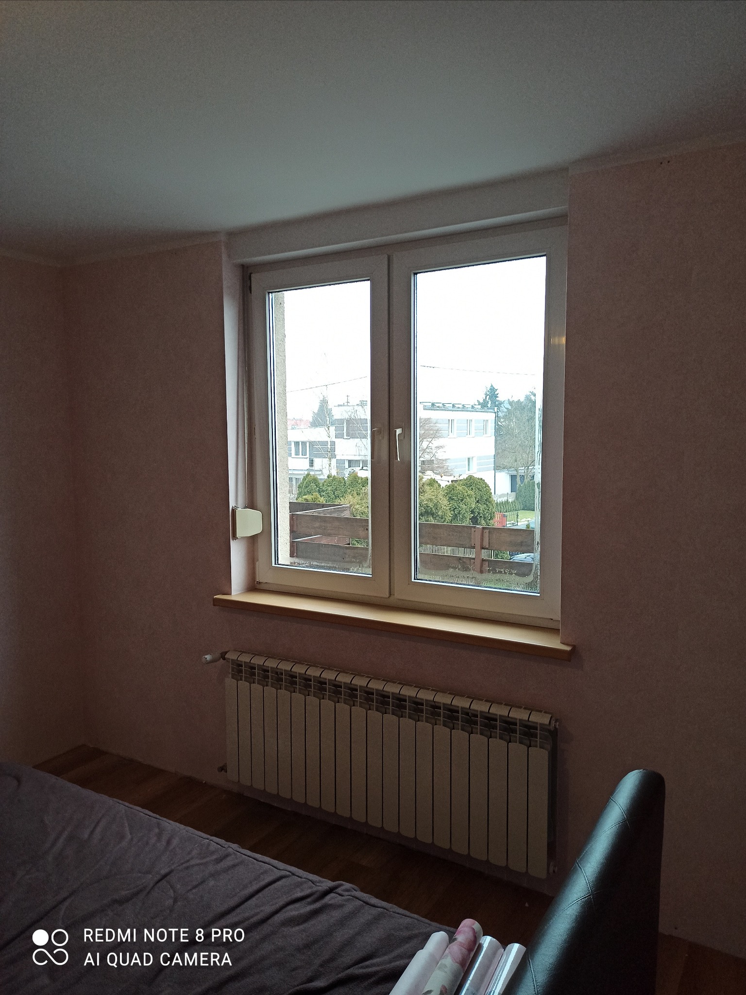 Pokój po odświeżeniu z widokiem na ogród przez okno, ściany pokryte jasnoróżową tapetą, pod oknem grzejnik, w rogu widoczne rolki tapet.