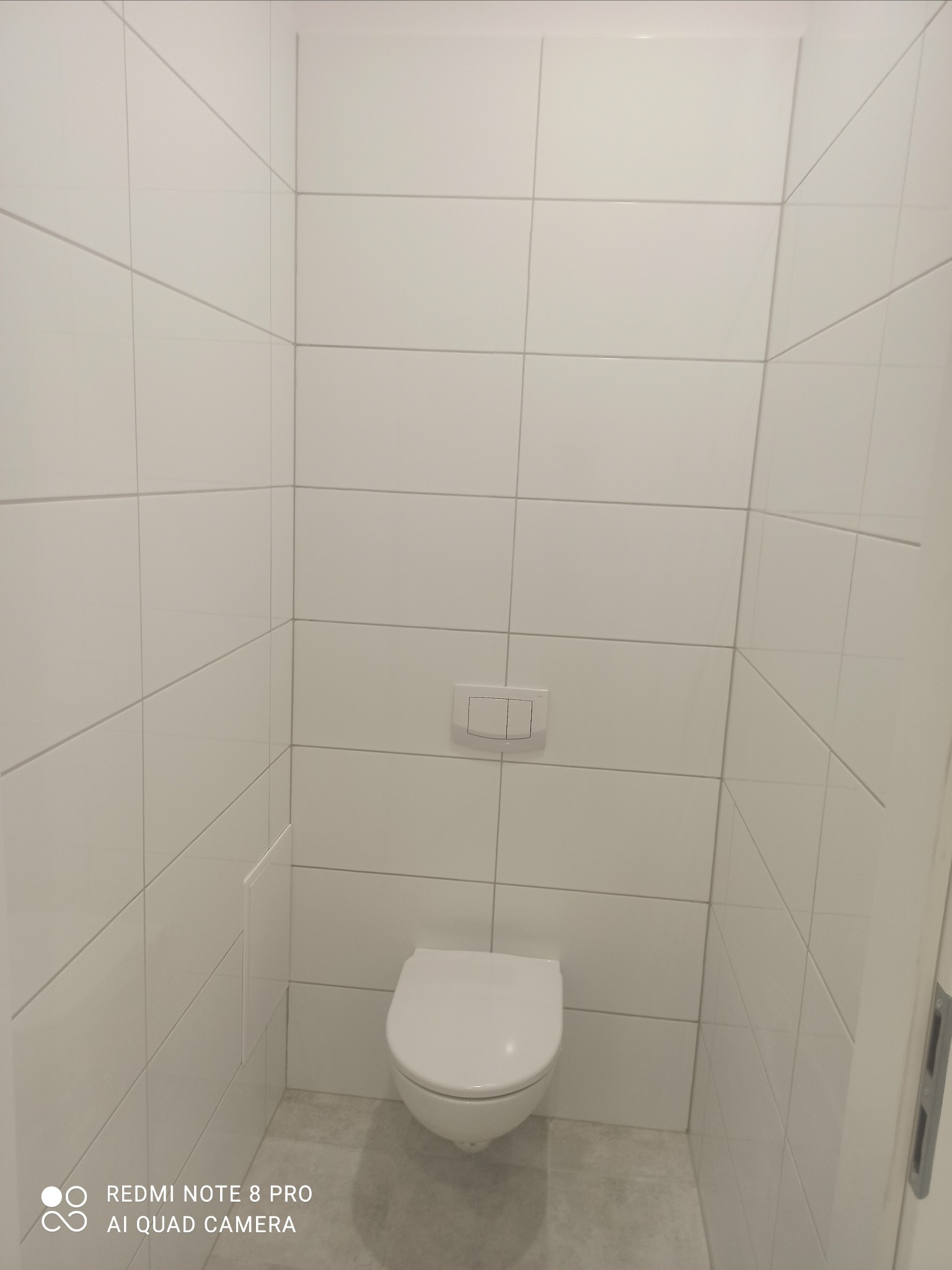 Wykończona toaleta z białymi, kwadratowymi płytkami na ścianach, podwieszana miska WC i szarymi fugami, widoczny fragment drzwi po prawej stronie.