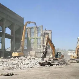 Trzy żółte koparki pracują nad rozbiórką częściowo zburzonego, wielopiętrowego budynku z betonu i cegły, z widocznymi oknami bez szyb i stertą gruzu na pierwszym planie.