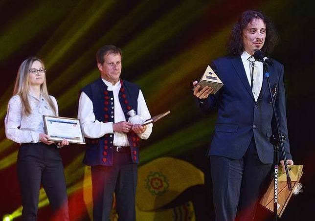 Trzech prelegentów na scenie podczas uroczystości wręczenia nagród, mężczyzna w garniturze trzyma trójkątną statuetkę, kobieta z blond włosami i okularach trzyma certyfikat, mężczyzna w ludowym...