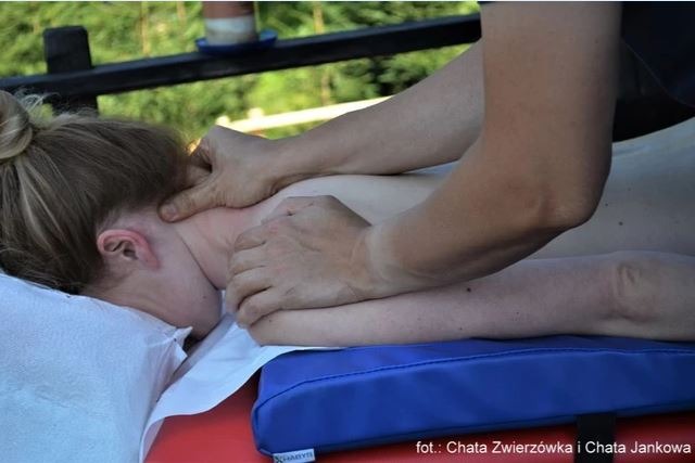 Pacjentka leżąca na leżance z niebieską poduszką pod klatką piersiową, podczas masażu szyi wykonywanego przez masażystę na świeżym powietrzu.