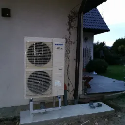 Zewnętrzna jednostka dwuwentylatorowa pompy ciepła Panasonic Inverter, zamontowana na betonowej podstawie przy ścianie domu z widocznym ogrodem w tle.