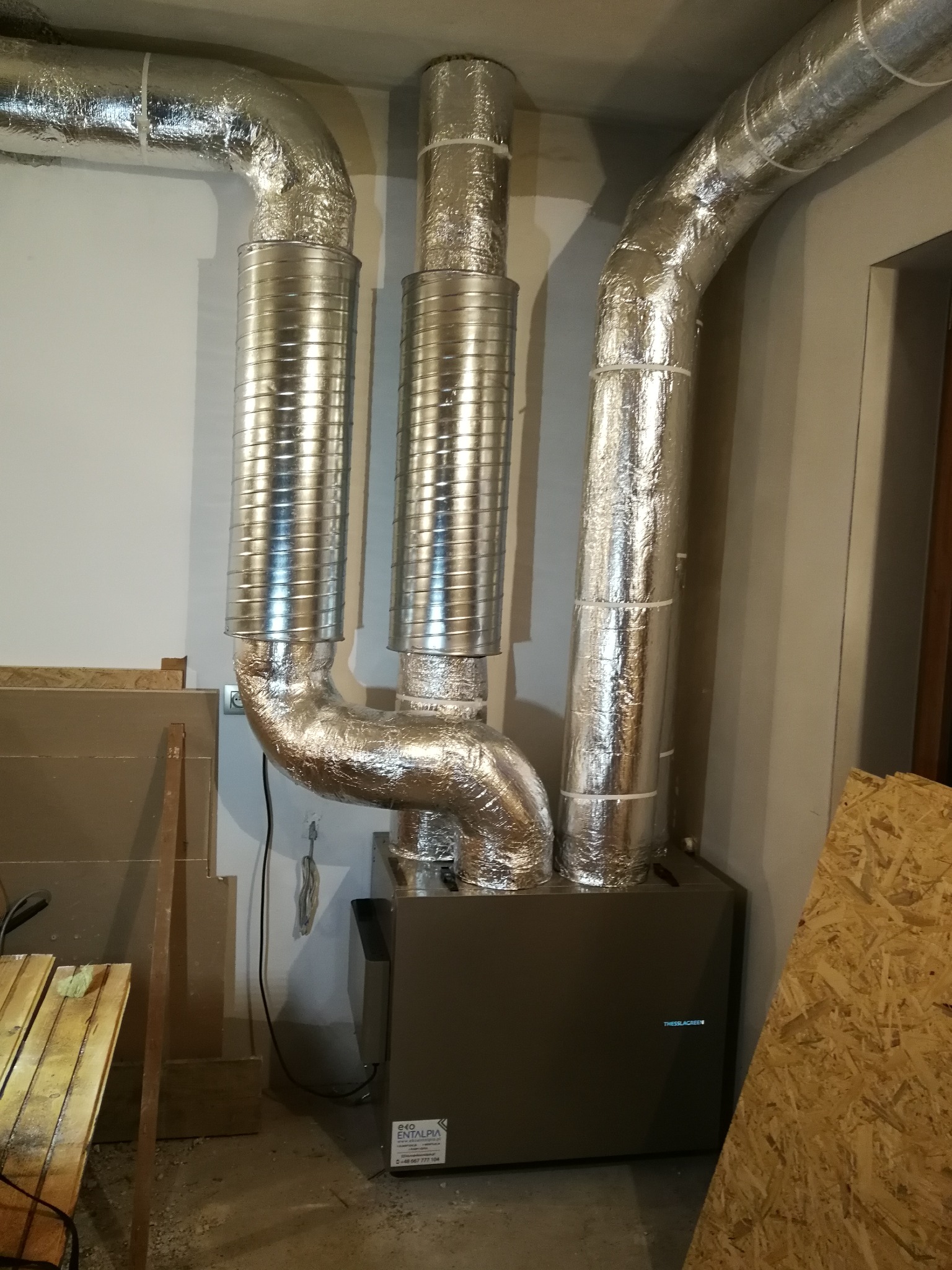 Instalacja rekuperacji z trzema przewodami wentylacyjnymi w srebrnej izolacji, widoczny fragment urządzenia z naklejką informacyjną