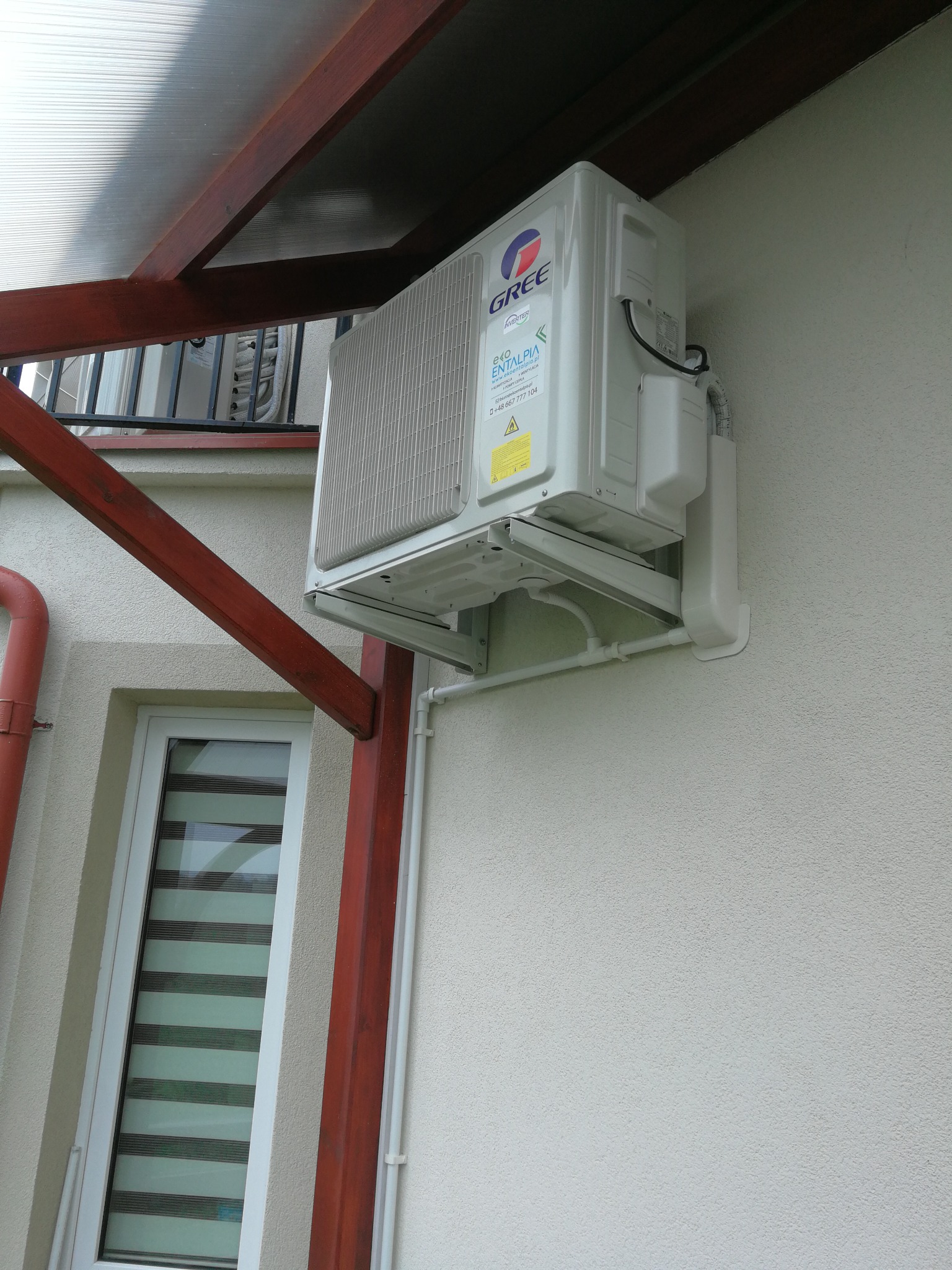 Zamontowana jednostka zewnętrzna klimatyzatora marki Gree na elewacji budynku z widocznym orynnowaniem i oknem z żaluzjami poziomymi.