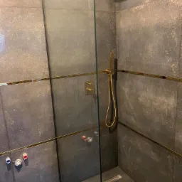 Szara kabina prysznicowa z elementami w kolorze złotym, widoczne podłączenia hydrauliczne do baterii podtynkowej.