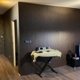 Firma ogólnobudowlana FACHDOM Jarosław Lewiński - Pokój po remoncie z ciemnobrązową ścianą imitującą drewno, fragment ściany z geometrycznym wzorem, telewizor z włączonym filmem i drewniana podłoga.