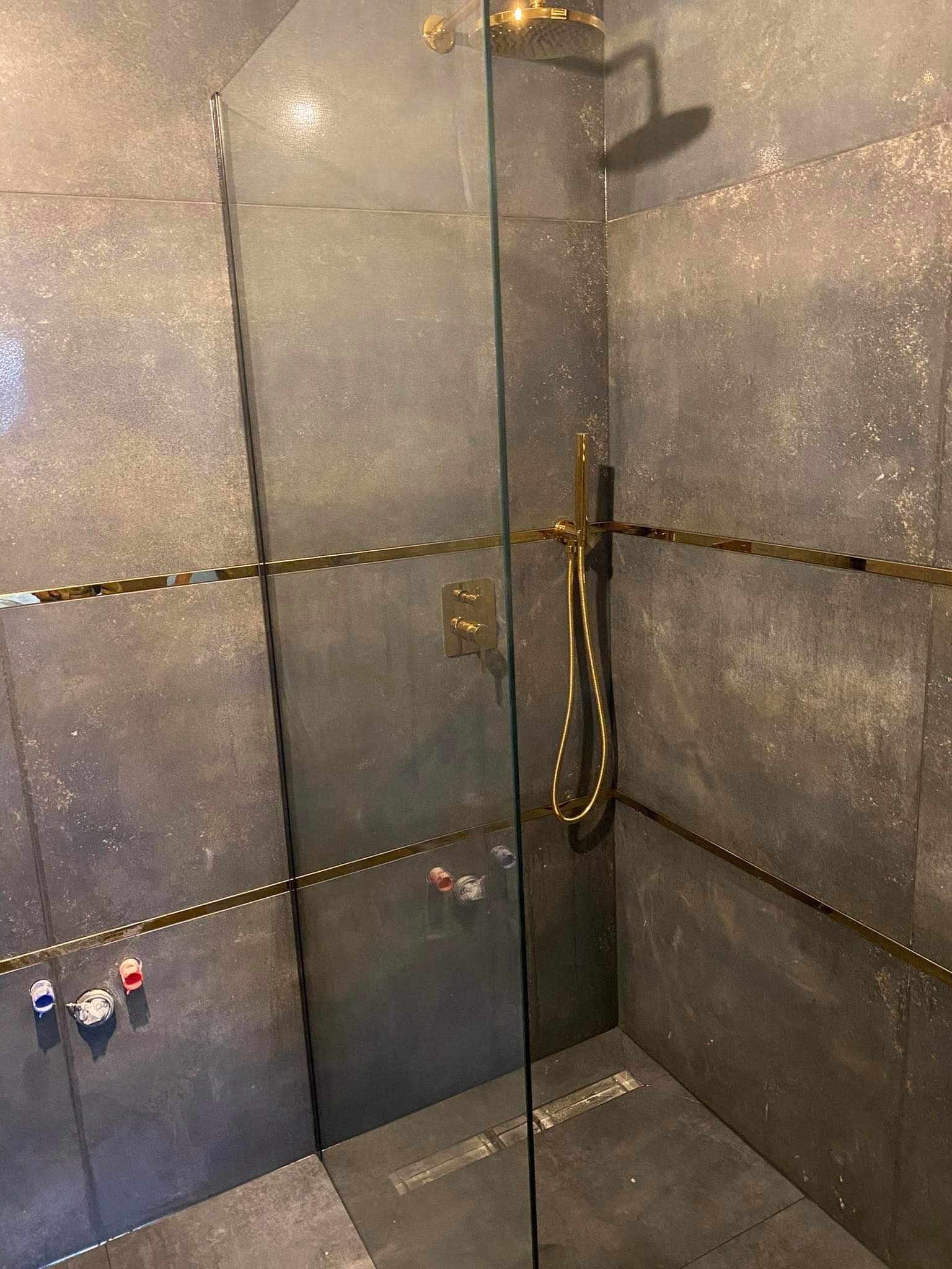 Szara kabina prysznicowa z elementami w kolorze złotym, widoczne podłączenia hydrauliczne do baterii podtynkowej.