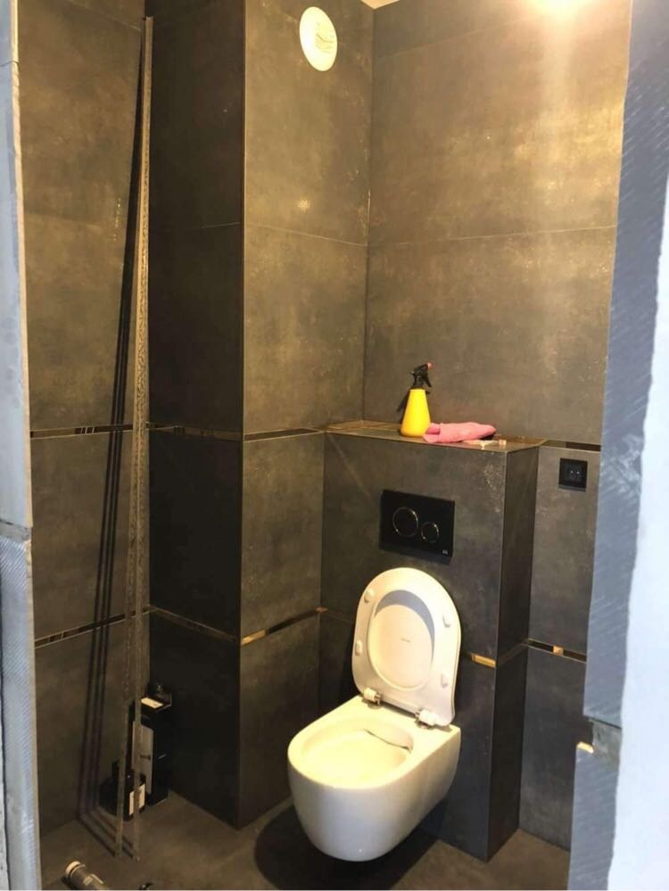 Nowoczesna toaleta podwieszana, obudowana ciemnymi płytkami z dekoracyjnymi złotymi paskami, żółty spryskiwacz i różowa ściereczka na obudowie stelaża.