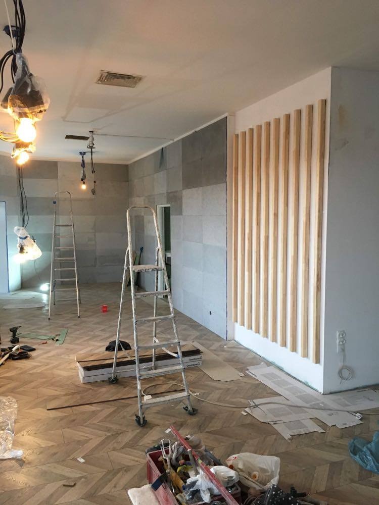 Wnętrze w trakcie prac: drewniane lamele na ścianie w pionowym układzie, surowe żarówki zwisające z sufitu, drabiny i narzędzia na parkiecie ułożonym we wzór jodełki.