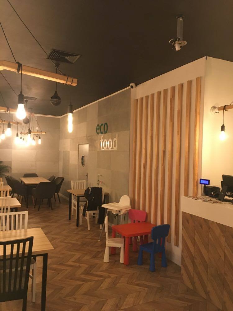 Wnętrze restauracji z napisem 'eco food' na ścianie, drewnianą podłogą w jodełkę i stolikami dla dzieci w kolorach czerwonym i niebieskim.