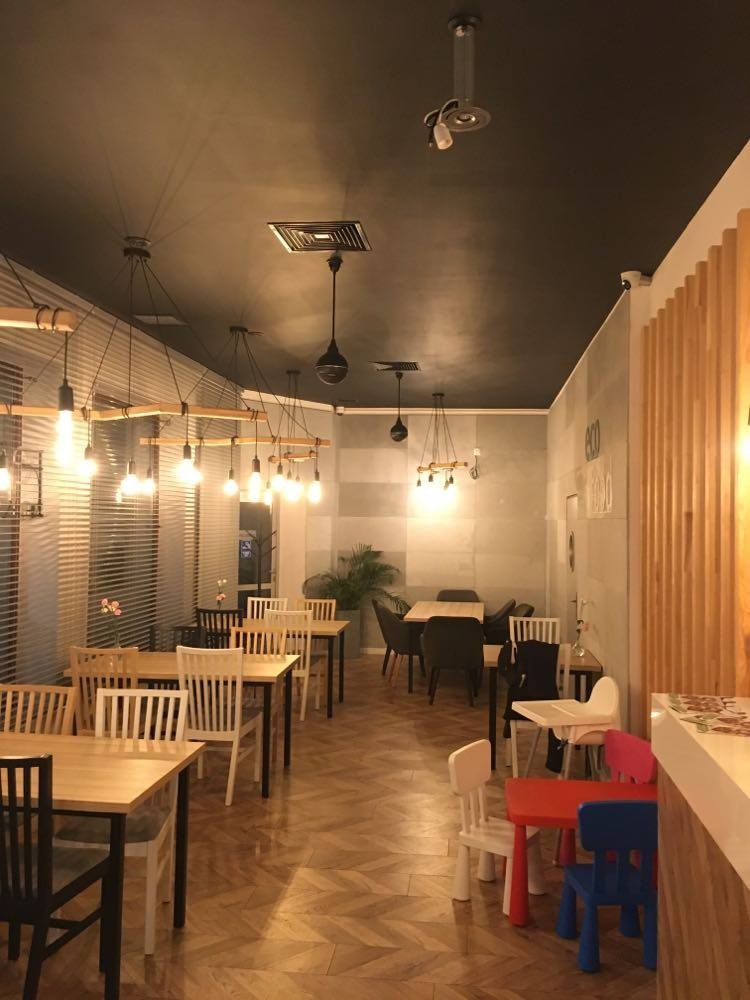 Wnętrze lokalu gastronomicznego z drewnianą podłogą w jodełkę, stołami i krzesłami o różnym designie, wiszącymi lampami z żarówkami Edison i czarnym sufitem z kratkami wentylacyjnymi.