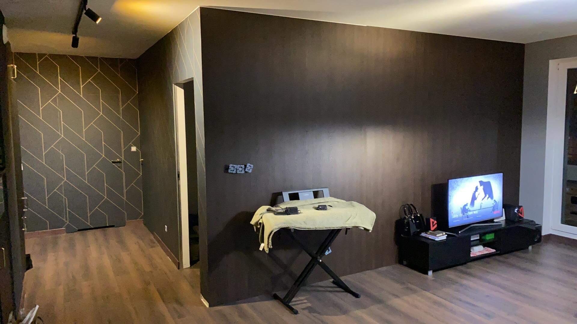 Pokój po remoncie z ciemnobrązową ścianą imitującą drewno, fragment ściany z geometrycznym wzorem, telewizor z włączonym filmem i drewniana podłoga.