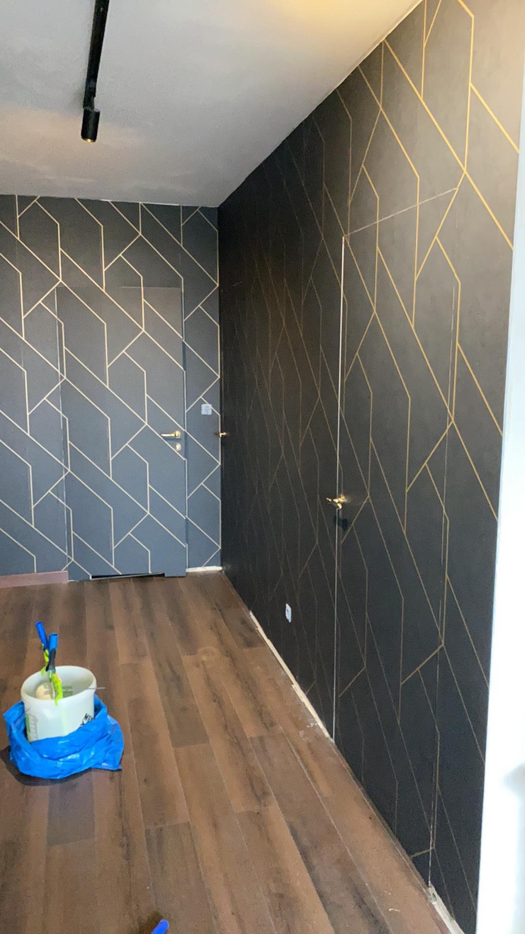 Wnętrze po odświeżeniu: ściany w ciemnoszarym kolorze z geometrycznym wzorem w odcieniach złota, drewniana podłoga, wiadro z narzędziami malarskimi w rogu pomieszczenia, drzwi ukryte w ścianie...