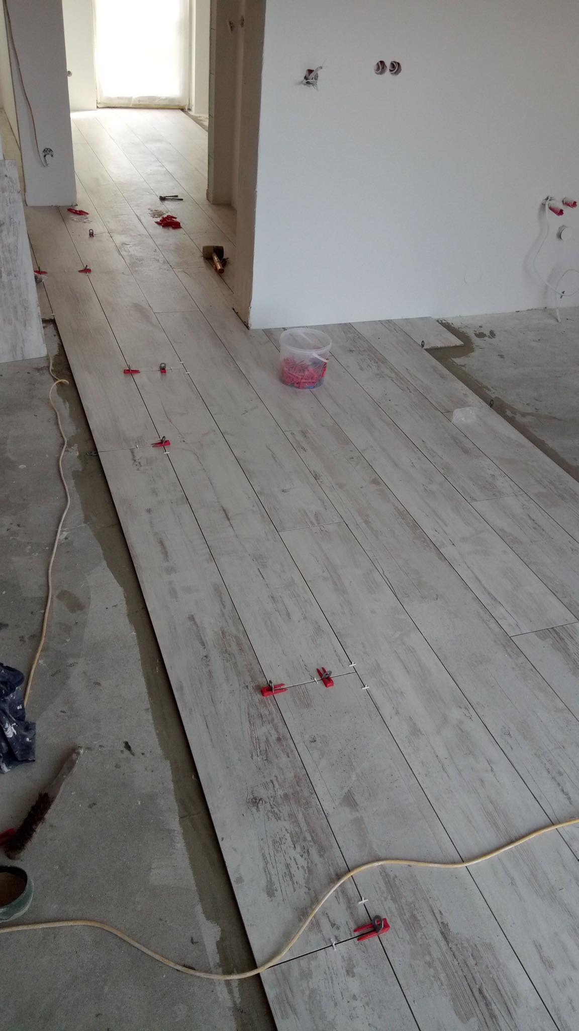 Układanie podłogi z płytek imitujących drewno, z użyciem klipsów poziomujących, widoczny kabel i wiadro z klinami poziomującymi w surowym wnętrzu.
