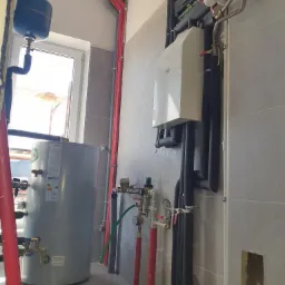 Nowoczesna kotłownia z szarym bojlerem, białym piecem i licznymi rurami w kolorze czerwonym i czarnym, zamontowana na szarej ścianie z płytek.