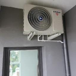 Zamontowana jednostka zewnętrzna klimatyzacji Kaisai na szarej ścianie balkonu, widoczne rurki odprowadzające skropliny i fragment okna z widokiem na zieleń.
