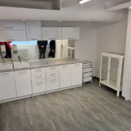 Wnętrze gabinetu lekarskiego z białymi szafkami, umywalkami i dużym lustrem, w którym odbijają się sylwetki osób.