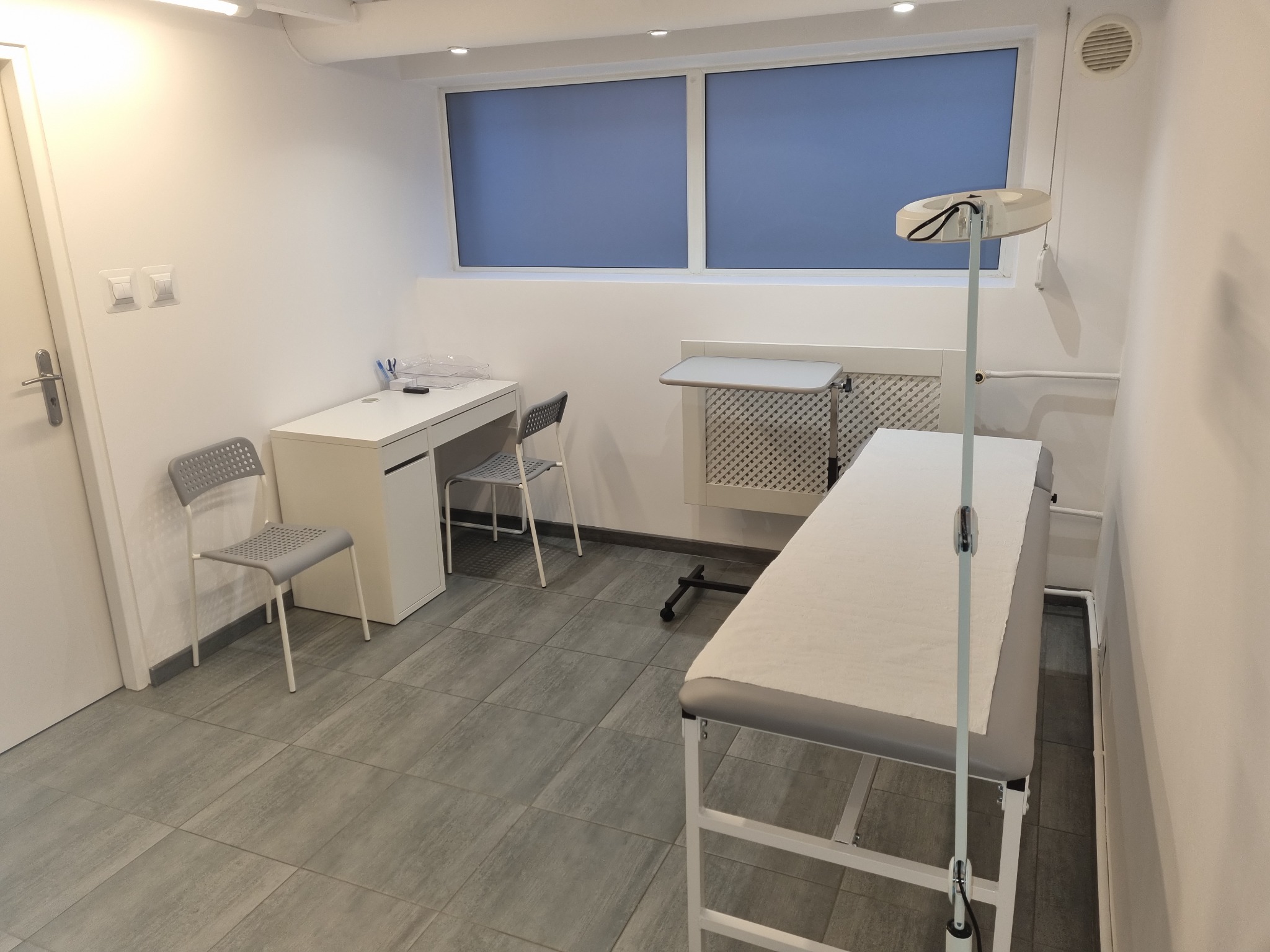 Jasne wnętrze gabinetu lekarskiego z kozetką pokrytą białym prześcieradłem, biurkiem, krzesłami i lampą medyczną