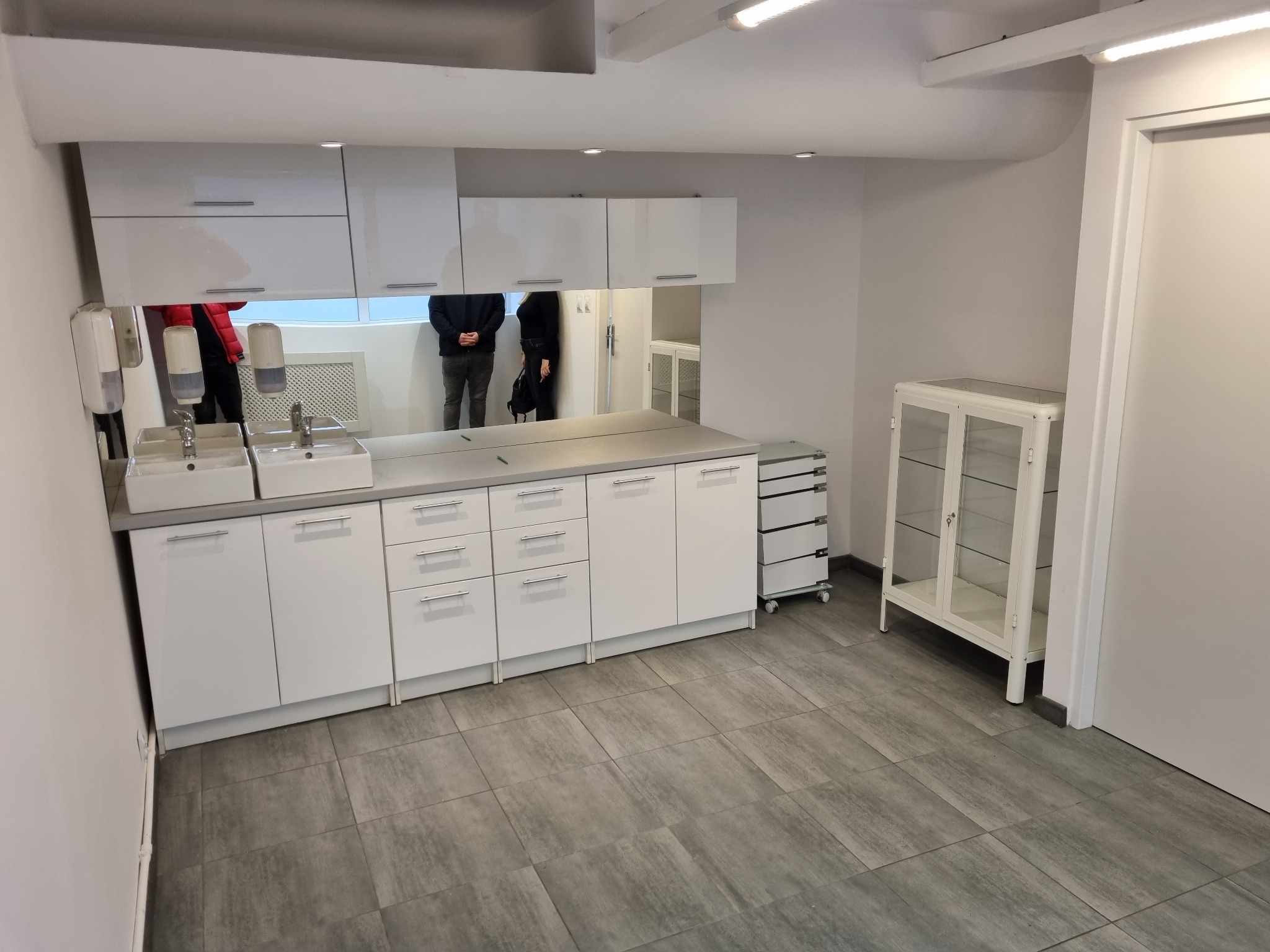 Wnętrze gabinetu lekarskiego z białymi szafkami, umywalkami i dużym lustrem, w którym odbijają się sylwetki osób.