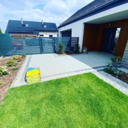 Nowoczesny dom jednorodzinny z szarą, poziomą bramą wjazdową i ogrodzeniem, jasną kostką brukową, zadbanym trawnikiem i rabatami, na których widać świeżo posadzone kwiaty. Na kostce leżą worki...