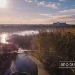 Panoramiczny widok z lotu ptaka na jezioro, las i odległe budynki przemysłowe w słoneczny dzień. Mały mostek z pojedynczą osobą przechodzącą przez niego przecina część jeziora. Logo w prawym dolnym...