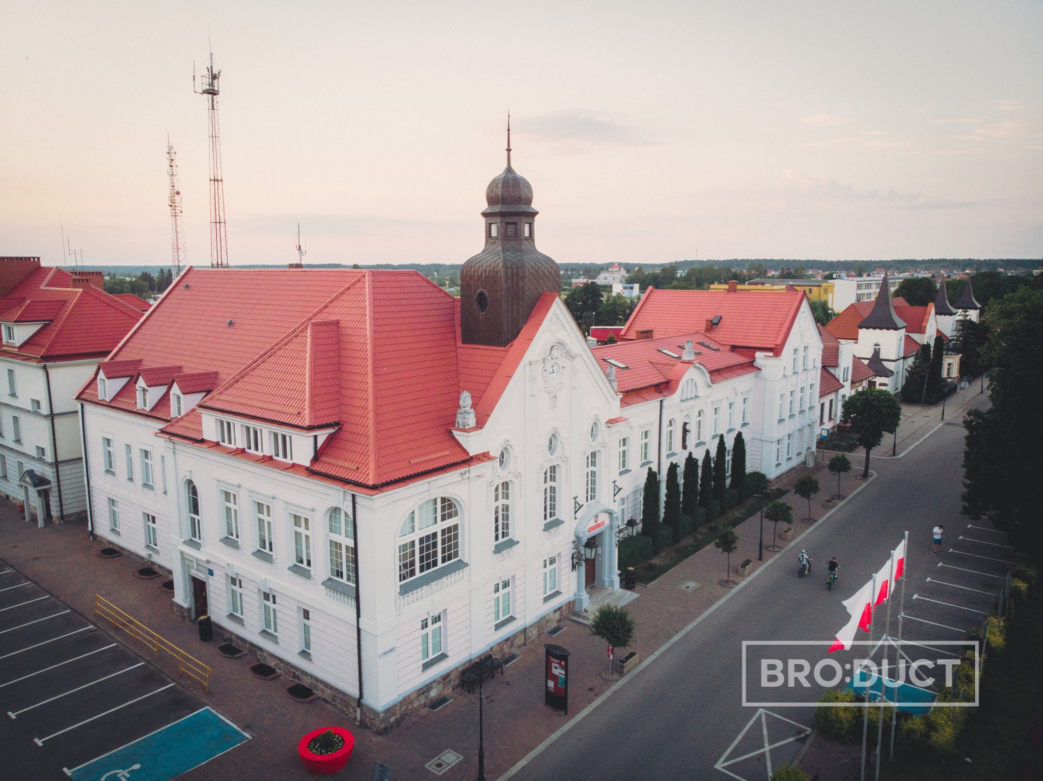 Widok z lotu ptaka na biały budynek z czerwoną dachówką, wieżyczką i parkingiem, z polskimi flagami na pierwszym planie i osobami na rowerach w tle.
