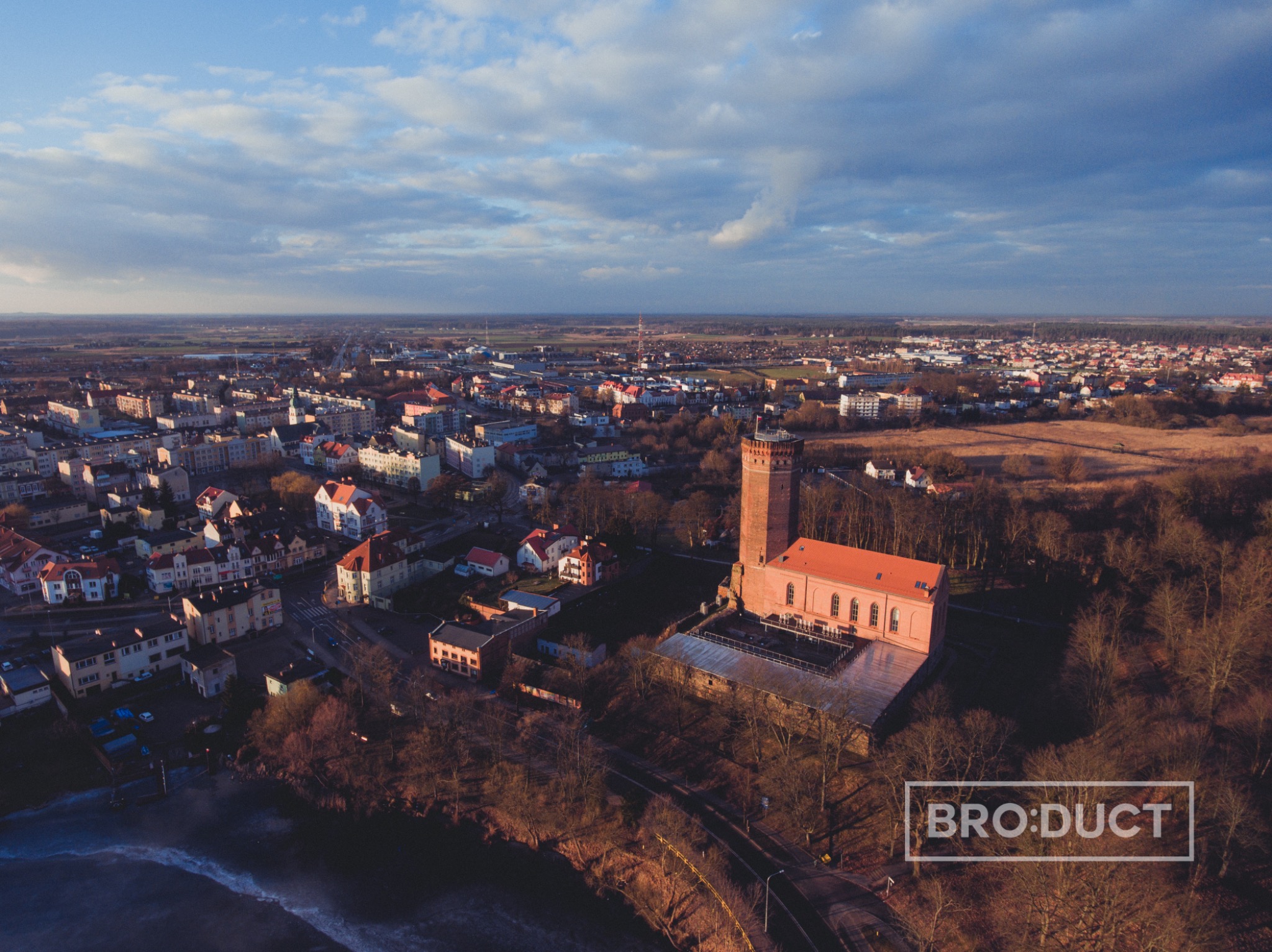 Panoramiczny widok z lotu ptaka na miasto z ceglaną wieżą kościelną i czerwonym dachem, widziany w promieniach zachodzącego słońca, z logo BRO:DUCT w prawym dolnym rogu.