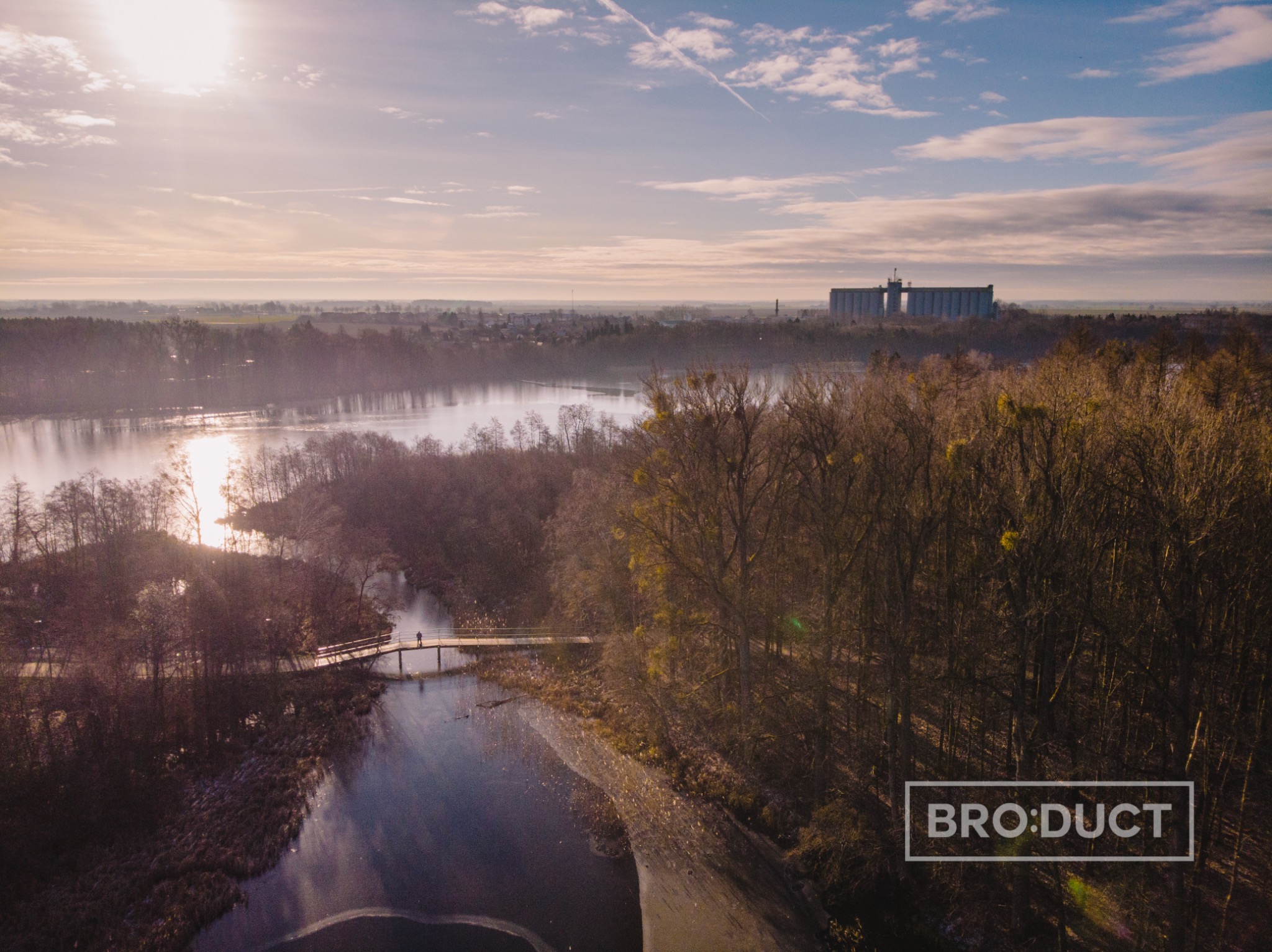 Panoramiczny widok z lotu ptaka na jezioro, las i odległe budynki przemysłowe w słoneczny dzień. Mały mostek z pojedynczą osobą przechodzącą przez niego przecina część jeziora. Logo w prawym dolnym...
