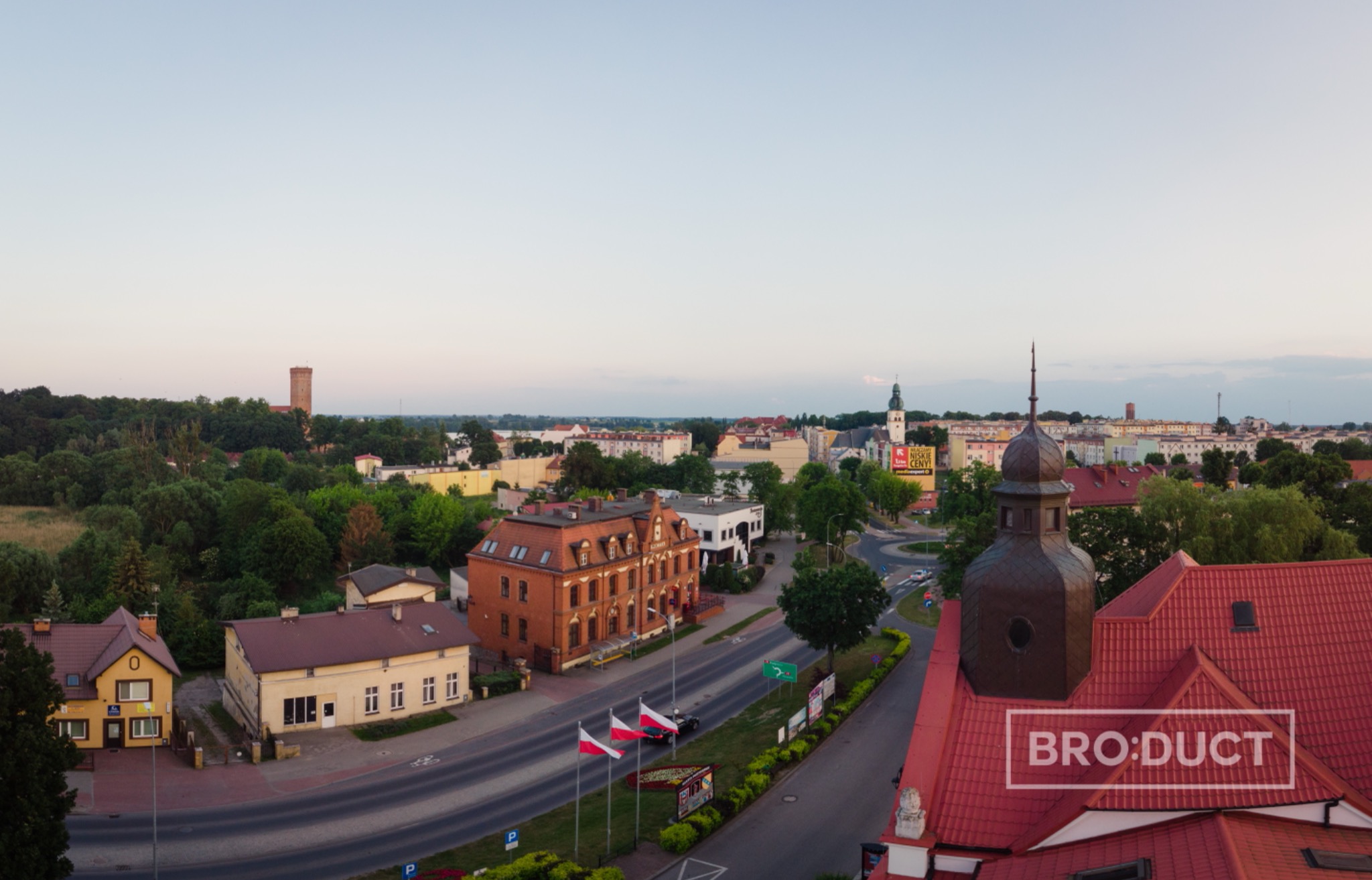 Panorama miasta z lotu ptaka o zmierzchu, widoczne budynki, drzewa, wieża ciśnień w tle, polskie flagi przy drodze.