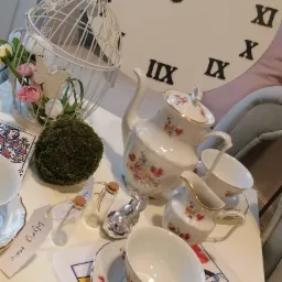 Dekoracja stołu w Bolesławcu z porcelanową zastawą w kwiaty, białą klatką dla ptaków z kwiatami i mchem, małymi szklanymi buteleczkami z korkami i figurką srebrnego zająca, na tle dekoracji...