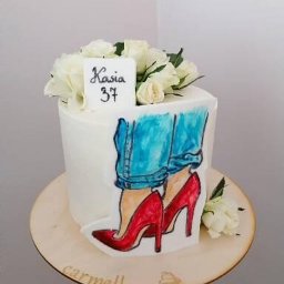 Pracownia Słodkości Carmellova - Tort urodzinowy z białym kremem, dekorowany rysunkiem dżinsów i czerwonych szpilek, z napisem 'Kasia 37' i białymi różami na górze, umieszczony na drewnianej podstawie z logo 'carmellova'.