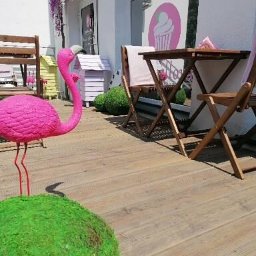 Pracownia Słodkości Carmellova - Zewnętrzny wystrój cukierni z różowym flamingiem, drewnianym stolikiem i krzesłami, dekoracjami kwiatowymi i logo babeczki na oknie.