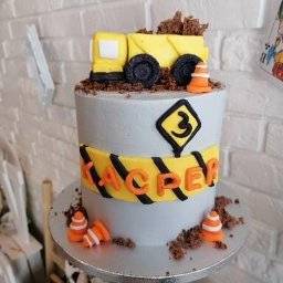 Pracownia Słodkości Carmellova - Tort urodzinowy w stylu budowlanym z żółtą ciężarówką na górze, dekorowany imieniem 'Kacper', cyfrą '3' i stożkami drogowymi na tle ceglanej ściany.