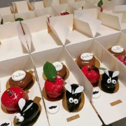 Zestaw unikalnych deserów w pudełkach z Bolesławca: błyszczące jabłka, czekoladowe ciastka z napisem 'tata' i owady z lukru, każdy na złotej podkładce.