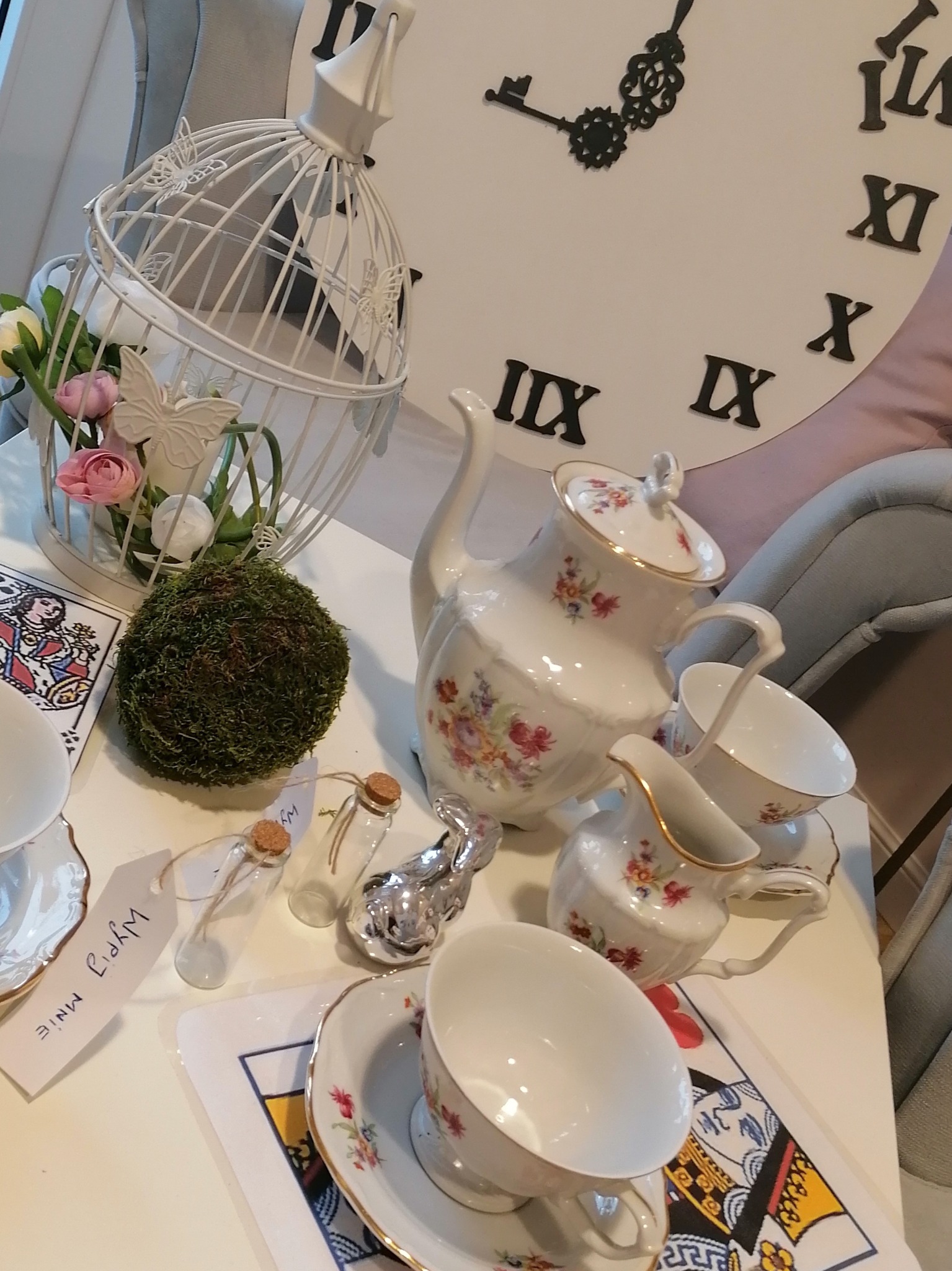 Dekoracja stołu w Bolesławcu z porcelanową zastawą w kwiaty, białą klatką dla ptaków z kwiatami i mchem, małymi szklanymi buteleczkami z korkami i figurką srebrnego zająca, na tle dekoracji...