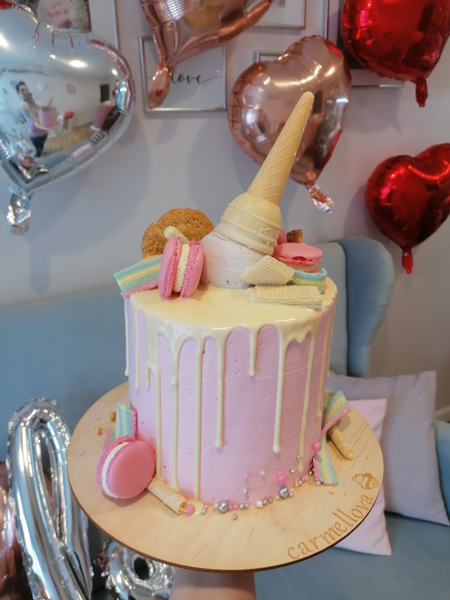 Różowy tort z polewą, dekorowany makaronikami, wafelkami, ciastkiem i rożkiem lodowym, na tle balonów w kształcie serc.