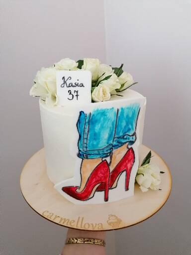 Tort urodzinowy z białym kremem, dekorowany rysunkiem dżinsów i czerwonych szpilek, z napisem 'Kasia 37' i białymi różami na górze, umieszczony na drewnianej podstawie z logo 'carmellova'.