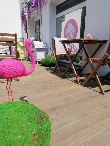 Zewnętrzny wystrój cukierni z różowym flamingiem, drewnianym stolikiem i krzesłami, dekoracjami kwiatowymi i logo babeczki na oknie.